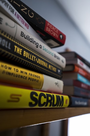 A stack of books is arranged on a wooden shelf, with titles facing outward. The books have varied colors and designs, with one prominently displaying the title 'SCRUM' in bold, yellow lettering. Others include 'Building a Second Brain' and works by 'Pierce Brown'. The setting is softly lit with a neutral background, suggesting a quiet and contemplative space.
