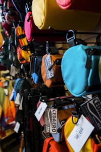 A display rack filled with a variety of colorful, small bags and pouches. The bags are made from fabric and come in colors such as yellow, red, blue, brown, and orange. Each bag has a label or tag attached to it, displaying the brand name 'CEDAERO' and indicating that the products are made in the USA. The bags are arranged in rows, hanging from hooks on a metal frame.