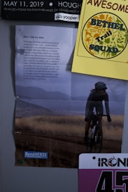 A collage of various items pinned on a wall, including a large poster at the center with text titled 'Why I ride my bike' and an image of a cyclist riding through a grassy landscape. Above it, a yellow paper with illustrations and the text 'BETHEL Trail SQUAD' alongside a calendar piece from May 11, 2019, showcasing event details. Part of a race bib with the number '457' is visible at the bottom right.