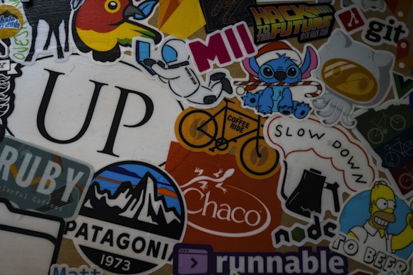 A colorful collage of various stickers featuring different designs, including a space-themed character, cartoon figures, a stylized mountain logo, coffee-themed graphics, and the word 'UP' in large font. There are references to popular culture, nature, and coding. Some stickers contain text, such as 'Hack to the Future,' 'Patagonia,' 'Coffee Ride,' and 'Slow Down.'