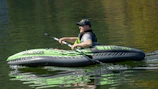A man wearing a dark cap, sunglasses, and a life jacket is paddling an inflatable kayak on a calm body of water. The kayak is green and gray, with the words 'CHALLENGER K1 INTEX' printed on the side.