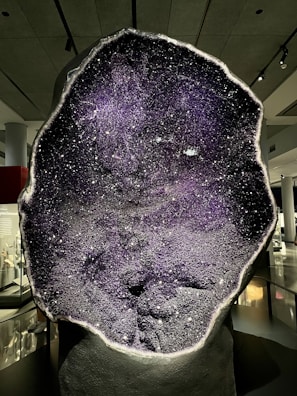 A large geode section reveals a sparkling, crystal-studded interior with a dominant purple hue. The geode is illuminated, enhancing the glittering effect of the crystals. It is displayed in a museum setting, with exhibits visible in the background under soft lighting.