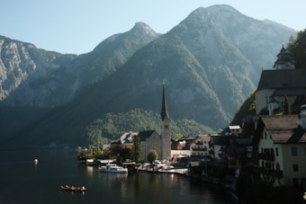 A picturesque village nestled along a serene lake with lush, green mountains towering in the background. The village architecture features charming, traditional buildings with steep roofs. A prominent church with a tall spire stands near the water's edge, while boats gently glide across the calm, reflective lake.