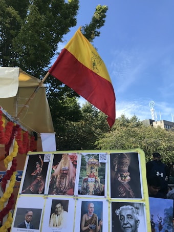 A vibrant scene featuring a flag with red and yellow stripes, prominently displayed against a bright blue sky. Below the flag is a collection of various portraits and images mounted on a board, including cultural and historical figures. The display is surrounded by yellow and red garlands, suggesting a festive or cultural event. In the background, lush green trees add to the lively atmosphere.