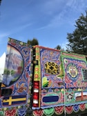 A vibrant and elaborately painted food truck with detailed artwork covering its surface. The designs include floral patterns, decorative elements, and a label reading 'FOOD TRUCK' in bold letters. There is also a sign with 'TRUCK WALL THE SPICY TRAILER' and a colorful mural featuring landscapes and intricate designs.