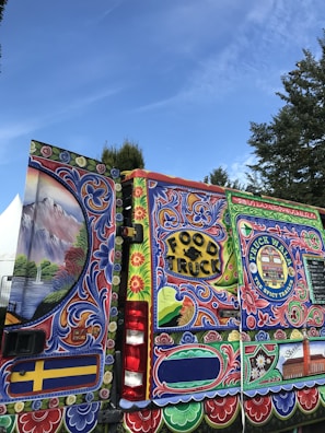 A vibrant and elaborately painted food truck with detailed artwork covering its surface. The designs include floral patterns, decorative elements, and a label reading 'FOOD TRUCK' in bold letters. There is also a sign with 'TRUCK WALL THE SPICY TRAILER' and a colorful mural featuring landscapes and intricate designs.