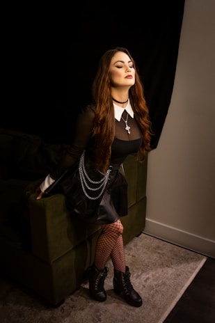 A person sits stylishly on the arm of a green sofa, wearing a black sheer top with a white collar, a cross necklace, a black skirt with chains, fishnet tights, and black lace-up boots. The setting has a dramatic and moody vibe, with low-key lighting against a dark backdrop.