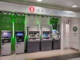 A row of five automated teller machines (ATMs) are stationed under a sign that reads 'Hang Seng Bank.' The machines have various interfaces and screens, with some displaying different languages and services. One of the machines has a sign indicating foreign exchange withdrawal. The area around the machines is clean and well-lit.