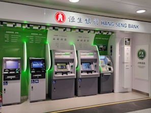 A row of five automated teller machines (ATMs) are stationed under a sign that reads 'Hang Seng Bank.' The machines have various interfaces and screens, with some displaying different languages and services. One of the machines has a sign indicating foreign exchange withdrawal. The area around the machines is clean and well-lit.