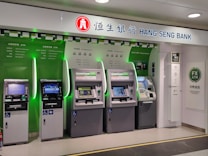 A row of five automated teller machines (ATMs) are stationed under a sign that reads 'Hang Seng Bank.' The machines have various interfaces and screens, with some displaying different languages and services. One of the machines has a sign indicating foreign exchange withdrawal. The area around the machines is clean and well-lit.
