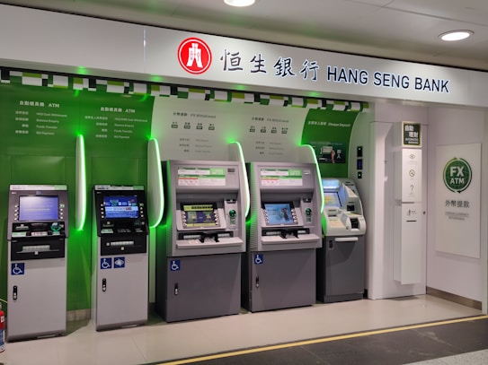A row of five automated teller machines (ATMs) are stationed under a sign that reads 'Hang Seng Bank.' The machines have various interfaces and screens, with some displaying different languages and services. One of the machines has a sign indicating foreign exchange withdrawal. The area around the machines is clean and well-lit.