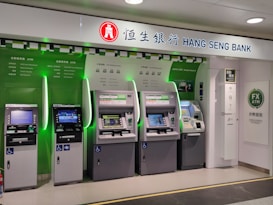 A row of five automated teller machines (ATMs) are stationed under a sign that reads 'Hang Seng Bank.' The machines have various interfaces and screens, with some displaying different languages and services. One of the machines has a sign indicating foreign exchange withdrawal. The area around the machines is clean and well-lit.