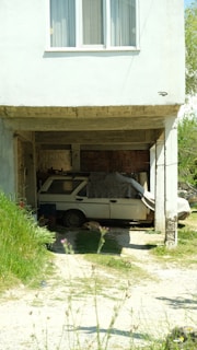 A white car is parked in an outdoor garage beneath a house. The car appears partially covered with a gray blanket or tarp. The garage space is open on one side, with brick walls visible in the background. The house above has a window with white curtains. The area surrounding the garage features some green grass and plants. There is also a dog lying on the ground near the car.