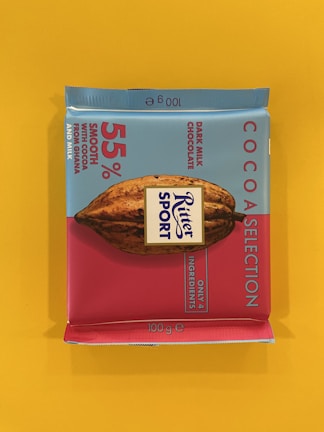 A square chocolate bar packaged in a colorful wrapper with a combination of light blue and pink colors. The branding prominently displays 'Ritter Sport' with details about the product, including '55% smooth with cocoa from Ghana and milk' and 'Dark Milk Chocolate'. A cocoa pod illustration is featured in the center.