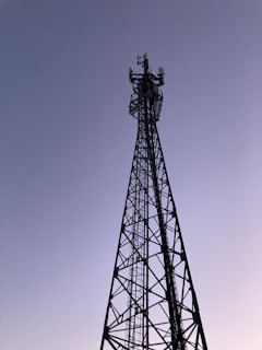 A tall metal communication tower reaches into a clear sky. The structure consists of a lattice framework, and there are several antennas mounted at the top. The sky has a gradient from light to dark blue, suggesting either early morning or late evening.