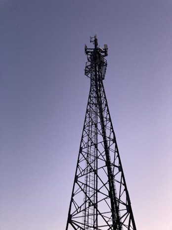 A tall metal communication tower reaches into a clear sky. The structure consists of a lattice framework, and there are several antennas mounted at the top. The sky has a gradient from light to dark blue, suggesting either early morning or late evening.