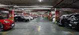 A spacious underground parking lot with numerous parked cars lined up along both sides of the aisle. The ceiling features exposed pipes and wiring, with red and white signage indicating parking rows and sections. The area is well-lit, creating a clean and organized atmosphere.