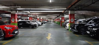 A spacious underground parking lot with numerous parked cars lined up along both sides of the aisle. The ceiling features exposed pipes and wiring, with red and white signage indicating parking rows and sections. The area is well-lit, creating a clean and organized atmosphere.