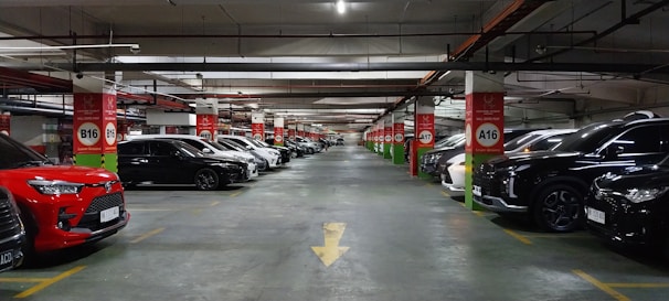 A spacious underground parking lot with numerous parked cars lined up along both sides of the aisle. The ceiling features exposed pipes and wiring, with red and white signage indicating parking rows and sections. The area is well-lit, creating a clean and organized atmosphere.
