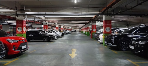 A spacious underground parking lot with numerous parked cars lined up along both sides of the aisle. The ceiling features exposed pipes and wiring, with red and white signage indicating parking rows and sections. The area is well-lit, creating a clean and organized atmosphere.
