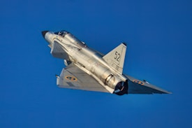 A jet aircraft with a sleek, silvery body is seen flying in a clear blue sky. The aircraft features delta wings and a distinctive tail design, with the number 52 and Swedish Air Force roundels visible on its wings.