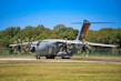 A large military transport aircraft is parked on a runway. The aircraft is gray with propellers in motion, indicated by the blurring. 'Luftwaffe' is written on the side, and 'Air Defender 2023' is visible on the tail. A backdrop of trees and a clear blue sky are in the background.
