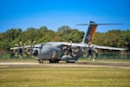 A large military transport aircraft is parked on a runway. The aircraft is gray with propellers in motion, indicated by the blurring. 'Luftwaffe' is written on the side, and 'Air Defender 2023' is visible on the tail. A backdrop of trees and a clear blue sky are in the background.