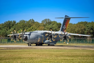 A large military transport aircraft is parked on a runway. The aircraft is gray with propellers in motion, indicated by the blurring. 'Luftwaffe' is written on the side, and 'Air Defender 2023' is visible on the tail. A backdrop of trees and a clear blue sky are in the background.