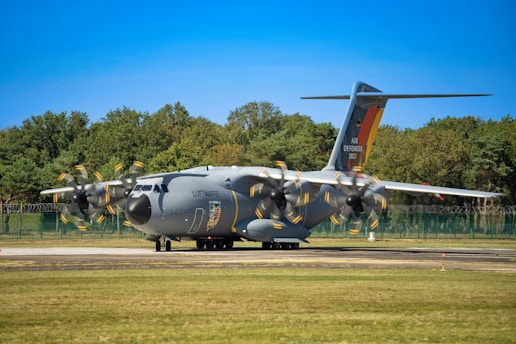 A large military transport aircraft is parked on a runway. The aircraft is gray with propellers in motion, indicated by the blurring. 'Luftwaffe' is written on the side, and 'Air Defender 2023' is visible on the tail. A backdrop of trees and a clear blue sky are in the background.