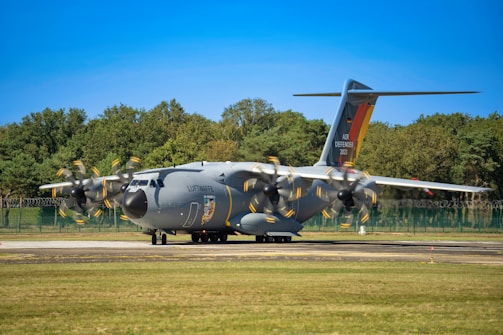 A large military transport aircraft is parked on a runway. The aircraft is gray with propellers in motion, indicated by the blurring. 'Luftwaffe' is written on the side, and 'Air Defender 2023' is visible on the tail. A backdrop of trees and a clear blue sky are in the background.
