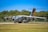 A large military transport aircraft is parked on a runway. The aircraft is gray with propellers in motion, indicated by the blurring. 'Luftwaffe' is written on the side, and 'Air Defender 2023' is visible on the tail. A backdrop of trees and a clear blue sky are in the background.