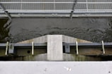 A metal footbridge runs across a water channel with visible flowing water. The bridge features a grid-like pattern on the walkway, supported by a sturdy metal frame. Below the bridge, there is a wooden and metal structure with bolts and beams.