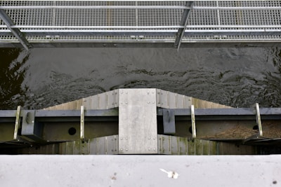 A metal footbridge runs across a water channel with visible flowing water. The bridge features a grid-like pattern on the walkway, supported by a sturdy metal frame. Below the bridge, there is a wooden and metal structure with bolts and beams.