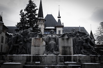 A dramatic stone sculpture features a central seated figure with outstretched arms, surrounded by additional human figures in dynamic poses. The backdrop is an elegant building with pointed towers and a historical architectural style. Inscriptions are visible on the stone base of the sculpture.