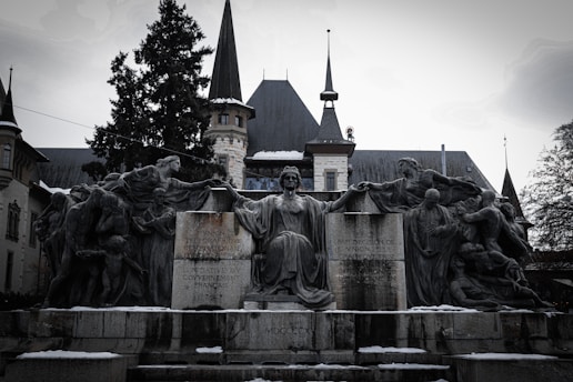 A dramatic stone sculpture features a central seated figure with outstretched arms, surrounded by additional human figures in dynamic poses. The backdrop is an elegant building with pointed towers and a historical architectural style. Inscriptions are visible on the stone base of the sculpture.
