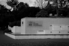 A black and white photograph features a building with a textured facade displaying the words 'Le Musée Olympique.' The structure is surrounded by trees and shrubs, creating a calm and serene setting.