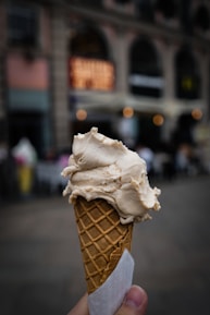 A hand holds a waffle cone with a scoop of creamy ice cream against an urban backdrop. The background is blurred, featuring a building with large windows and warm lights creating a cozy atmosphere.