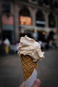A hand holds a waffle cone with a scoop of creamy ice cream against an urban backdrop. The background is blurred, featuring a building with large windows and warm lights creating a cozy atmosphere.