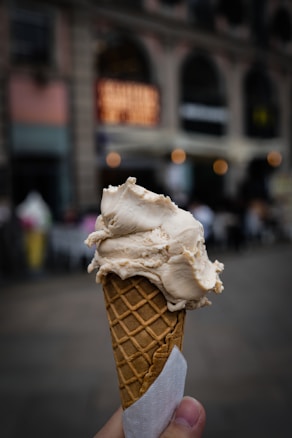 A hand holds a waffle cone with a scoop of creamy ice cream against an urban backdrop. The background is blurred, featuring a building with large windows and warm lights creating a cozy atmosphere.