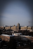A large, historic building with a dome and classical architecture dominates the scene. Surrounding the building are several smaller structures and snow-dusted rooftops, with cloudy skies overhead. Construction cranes are visible in the background, indicating development in the area.