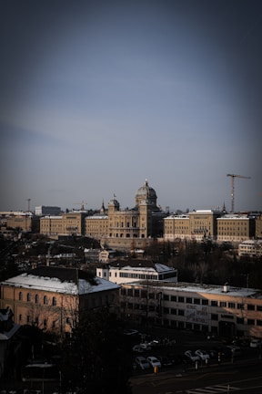 A large, historic building with a dome and classical architecture dominates the scene. Surrounding the building are several smaller structures and snow-dusted rooftops, with cloudy skies overhead. Construction cranes are visible in the background, indicating development in the area.