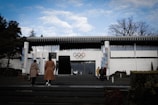 A modern building with a white facade and distinctive architecture, featuring the Olympic rings prominently displayed above the entrance. Several people are approaching the building, walking up steps from a landscaped area with trees and shrubs. The sky overhead is partly cloudy, adding a calm atmosphere.