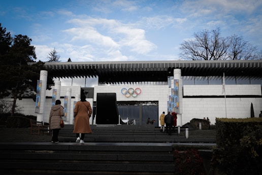 A modern building with a white facade and distinctive architecture, featuring the Olympic rings prominently displayed above the entrance. Several people are approaching the building, walking up steps from a landscaped area with trees and shrubs. The sky overhead is partly cloudy, adding a calm atmosphere.