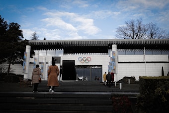A modern building with a white facade and distinctive architecture, featuring the Olympic rings prominently displayed above the entrance. Several people are approaching the building, walking up steps from a landscaped area with trees and shrubs. The sky overhead is partly cloudy, adding a calm atmosphere.