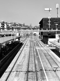 A monochrome image of a railway station with tracks stretching into the distance, flanked by platforms and modern buildings. Overhead wires and signal equipment are visible, contributing to an industrial and structured setting. The backdrop consists of low hills and scattered buildings under a clear sky.