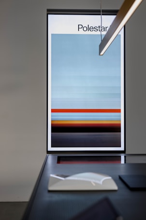 A large framed artwork with a gradient pattern of horizontal stripes in shades of blue, red, and black. It is displayed on a wall next to a modern table with a book and a minimalistic light fixture overhead.