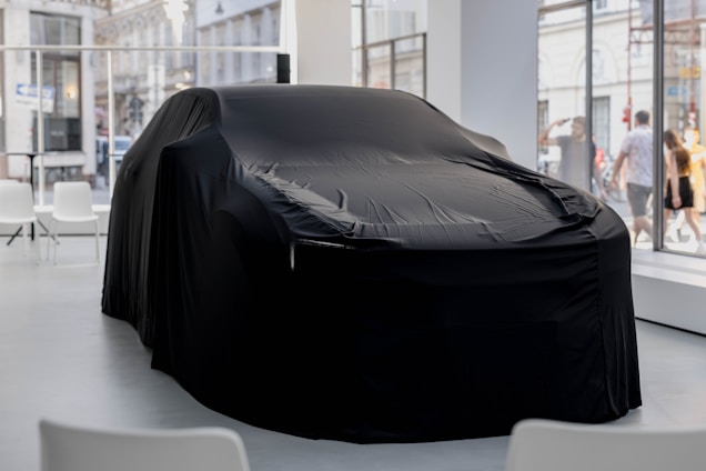 A covered car is centrally placed in a modern showroom with large windows. The black fabric draped over the vehicle conceals its shape, adding a sense of mystery. Chairs are positioned around the space, creating an atmosphere of anticipation. Outside, people are visible through the glass, walking past the showroom.