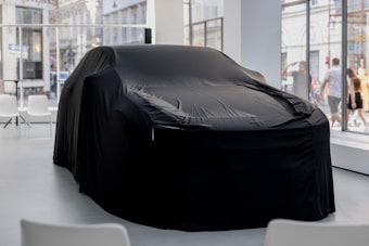 A covered car is centrally placed in a modern showroom with large windows. The black fabric draped over the vehicle conceals its shape, adding a sense of mystery. Chairs are positioned around the space, creating an atmosphere of anticipation. Outside, people are visible through the glass, walking past the showroom.