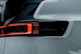 A close-up view of a car's rear light assembly featuring sleek, modern design. The light is illuminated with a bright red LED strip surrounding a dark, glossy housing. The word 'Polestar' is visible, suggesting the brand of the vehicle. The surrounding car body has a smooth, glossy white finish.