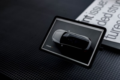 A tablet displays an image of a white car viewed from above. It is positioned on a dark, grid-patterned surface next to a stack of publications with black and white text partially visible.